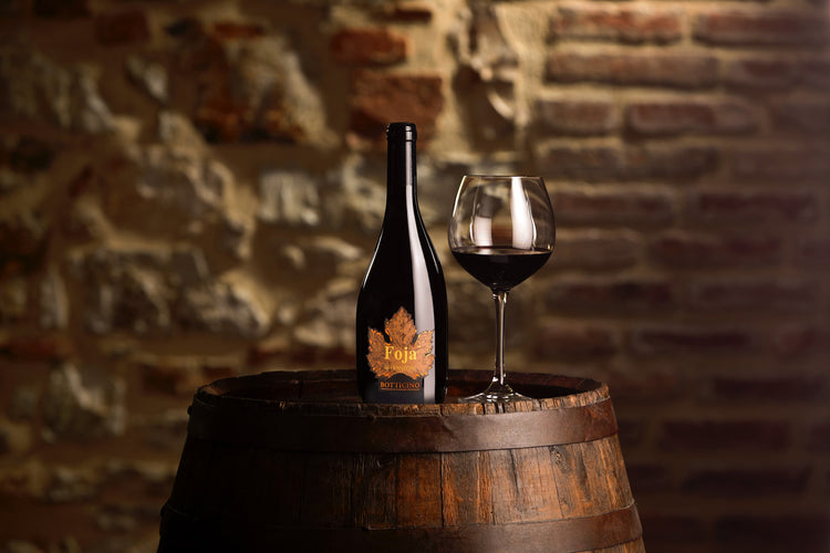 Bottiglia di Foja accanto a un calice di vino rosso su una botte in cantina
